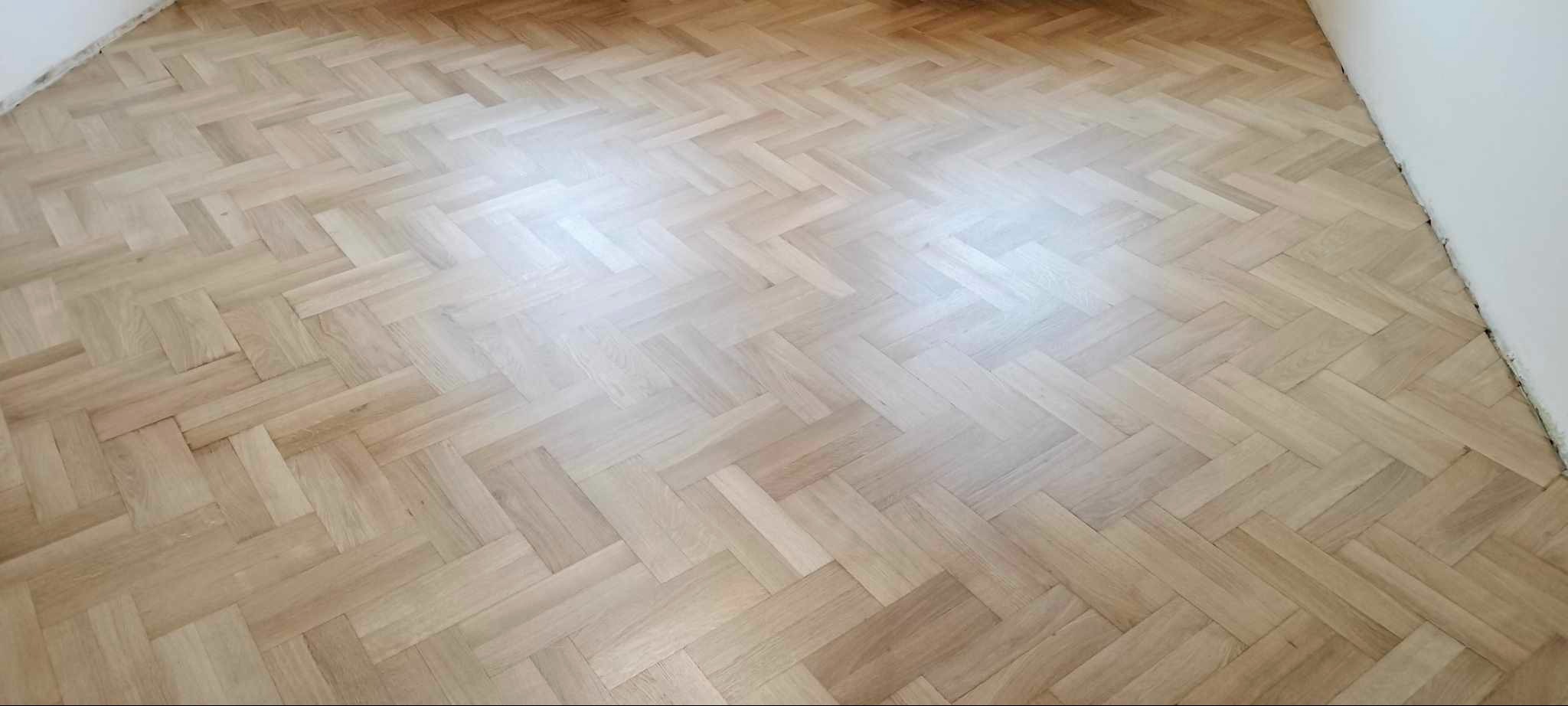 Jasny parkiet ułożony we wzór jodełki francuskiej, widok z góry, delikatne oświetlenie podkreśla naturalne usłojenie drewna, świeżo po renowacji.