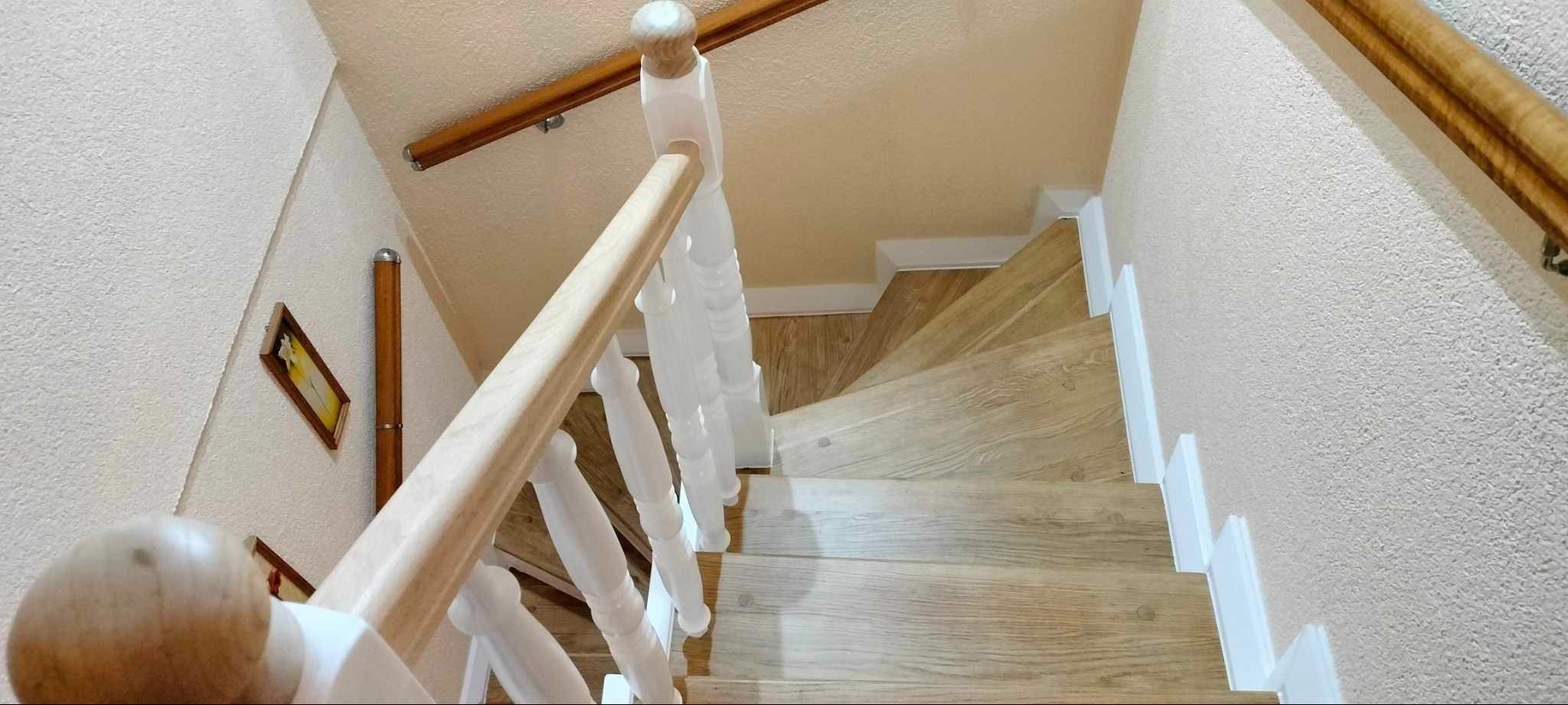 Jasne drewniane schody z białą balustradą, widok z góry. Na ścianie wisi obrazek w ramce. Naturalne światło podkreśla teksturę drewna i detale wykończenia.