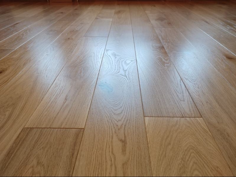 Jasny, lakierowany parkiet dębowy ułożony w regularny wzór. Widoczne naturalne usłojenie drewna i delikatne refleksy światła. Podłoga po cyklinowaniu i lakierowaniu.