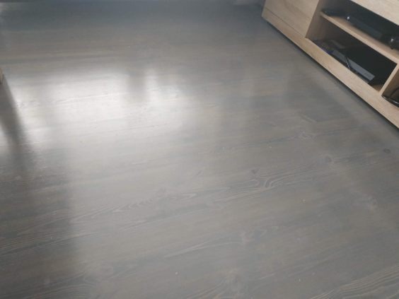 Gładka, szara drewniana podłoga po renowacji, z widocznym rysunkiem słojów, sąsiadująca z jasnym regałem na sprzęt RTV. Minimalistyczny design.