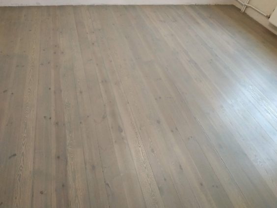 Jasnoszara, odnowiona podłoga z desek, widoczna pod kątem. Świeżo położona, z widoczną strukturą drewna i subtelnymi detalami wykończenia.