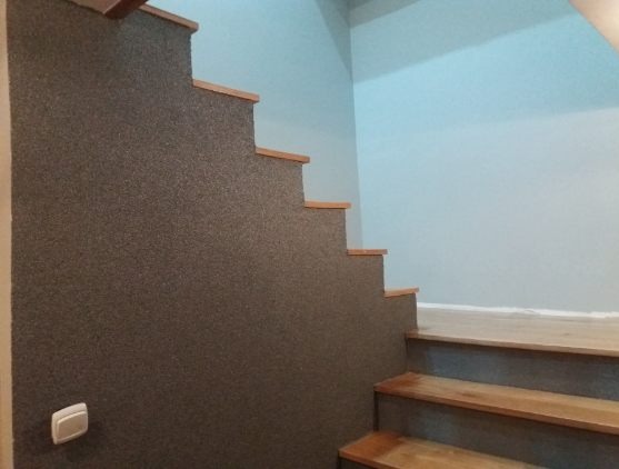 Schody z drewnianymi stopniami i ścianą pokrytą dekoracyjnym tynkiem w odcieniach szarości, oświetlone naturalnym światłem. Widoczny fragment ściany pomalowanej na jasny błękit.