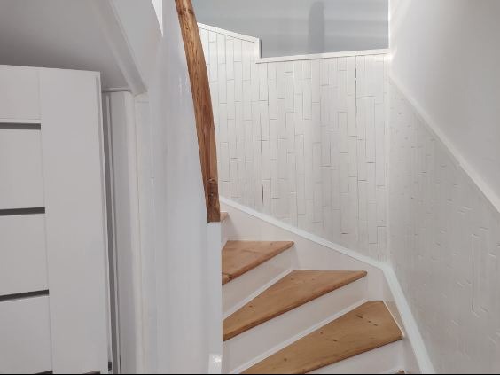 Jasne drewniane schody z białą balustradą i ścianami, widoczne z dołu. Minimalistyczny design z akcentem na naturalne materiały i proste linie.