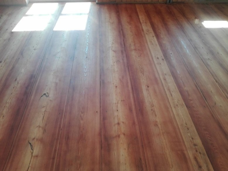 Wykończona podłoga z desek o ciepłym, czerwono-brązowym odcieniu, oświetlona naturalnym światłem wpadającym przez okno. Widoczne słoje drewna i subtelne niedoskonałości.