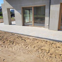 SEBRUK Sebastian Styczyński - Jasnoszara kostka brukowa ułożona przy nowoczesnym domu z dużymi oknami, widoczny piasek i ślady opon, sugerujące prace wykończeniowe.