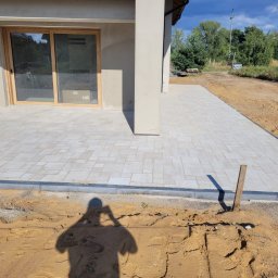 SEBRUK Sebastian Styczyński - Jasnoszara kostka brukowa ułożona na posesji, widoczny fragment domu z oknem i kolumną. W tle drzewa i niebo. W dolnej części zdjęcia ziemia i cień osoby.