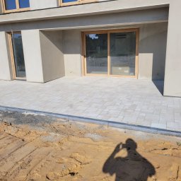 SEBRUK Sebastian Styczyński - Jasna kostka brukowa ułożona przed budynkiem z drewnianą stolarką okienną. W tle widoczna ziemia i cień osoby robiącej zdjęcie.