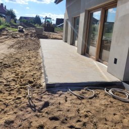 SEBRUK Sebastian Styczyński - Jasna kostka brukowa ułożona przy budynku z dużymi oknami, w tle ziemia i maszyna budowlana. Widok na częściowo ukończony taras.