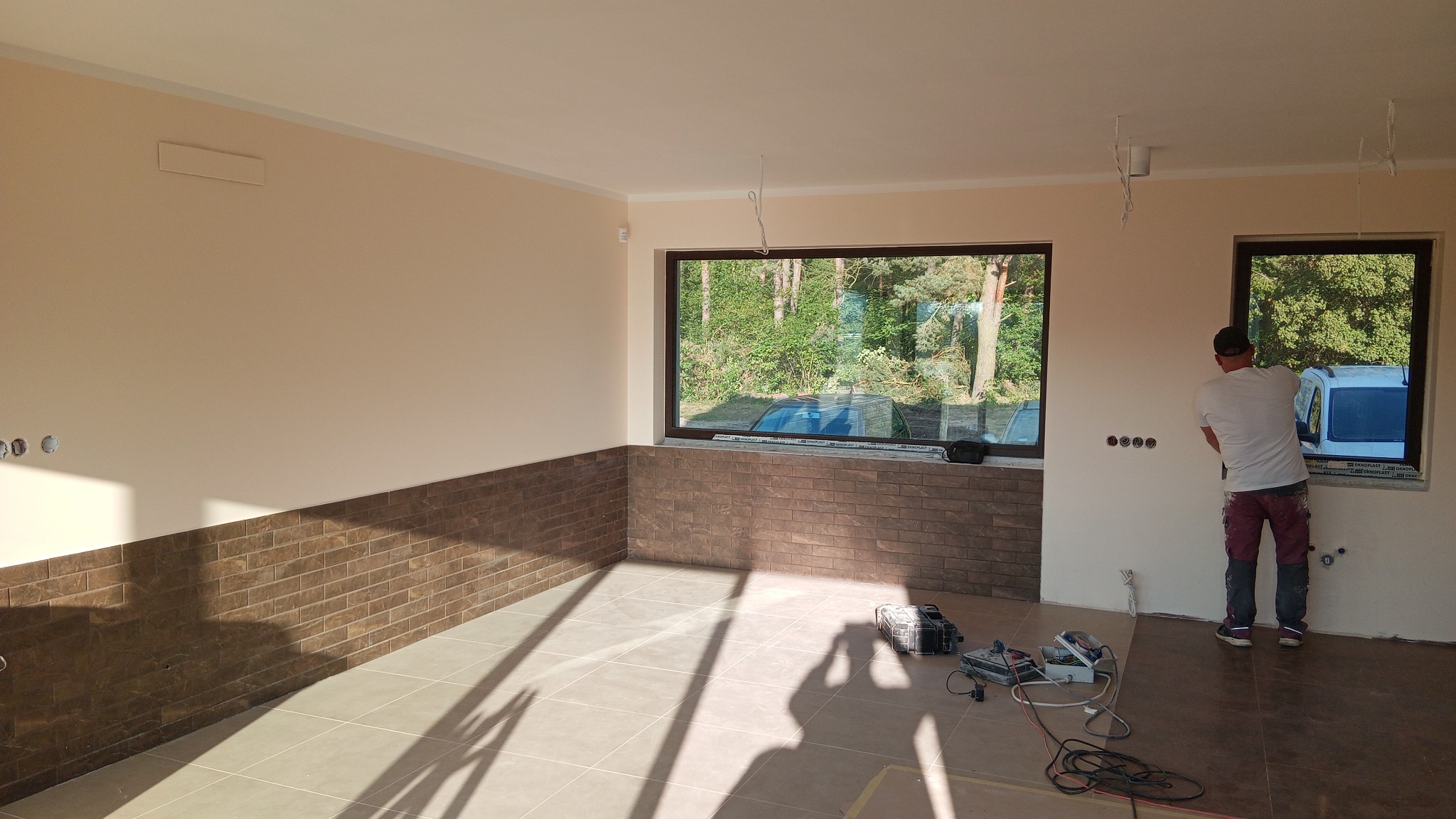 Wnętrze w trakcie wykańczania: częściowo wyłożona cegłą ściana, okna z widokiem na zieleń, pracownik przy oknie, narzędzia na podłodze. Jasne płytki podłogowe.