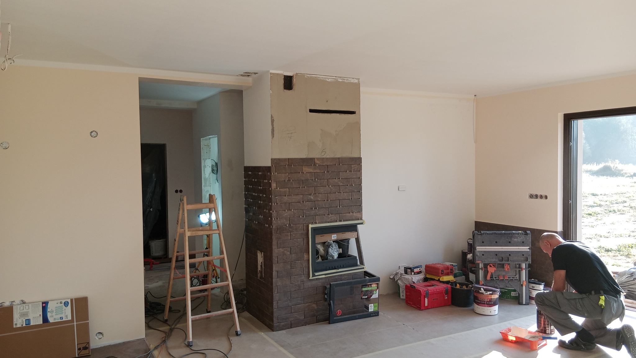 Wnętrze w trakcie wykańczania: częściowo obłożony brązową cegłą kominek, drabina, narzędzia i pracownik w trakcie pracy. Widoczne przygotowania do dalszych prac wykończeniowych.