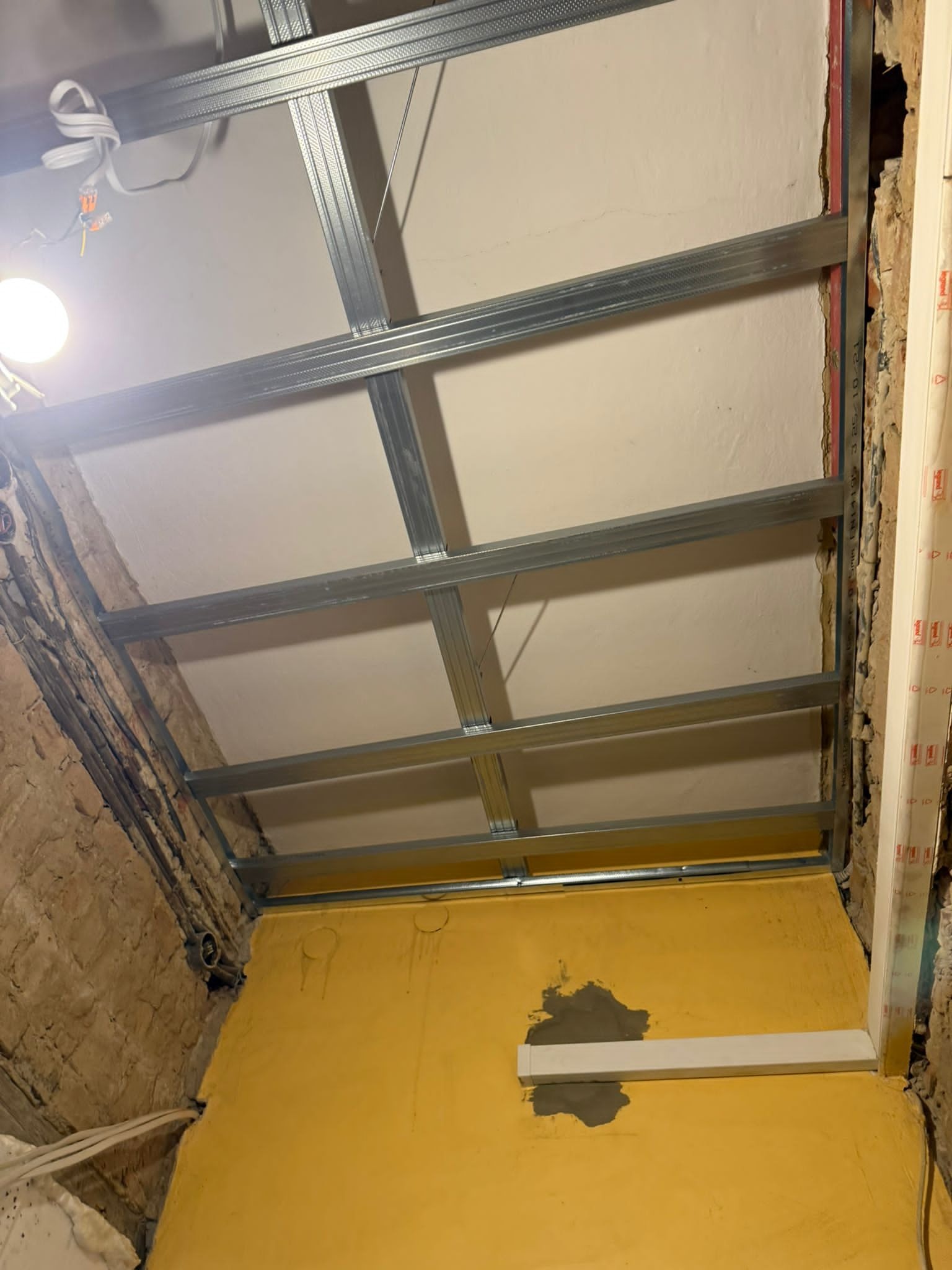 Montaż metalowej konstrukcji sufitu podwieszanego w pomieszczeniu z żółtą ścianą i odsłoniętą cegłą. Widoczne profile i instalacja elektryczna.