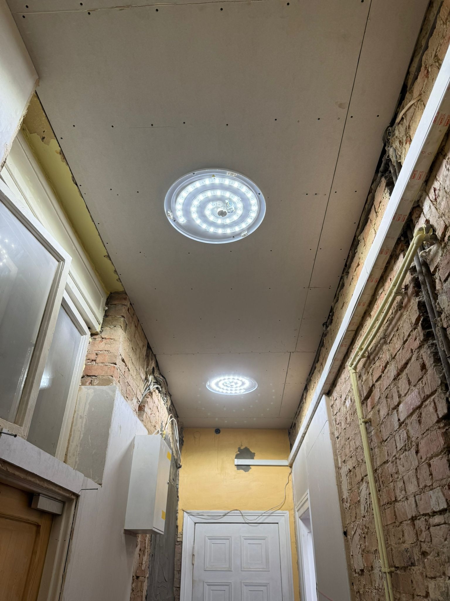 Montaż sufitu podwieszanego z płyt gipsowych i oświetleniem LED w korytarzu z odsłoniętą cegłą. Widoczne instalacje elektryczne i rury. Etap prac wykończeniowych.