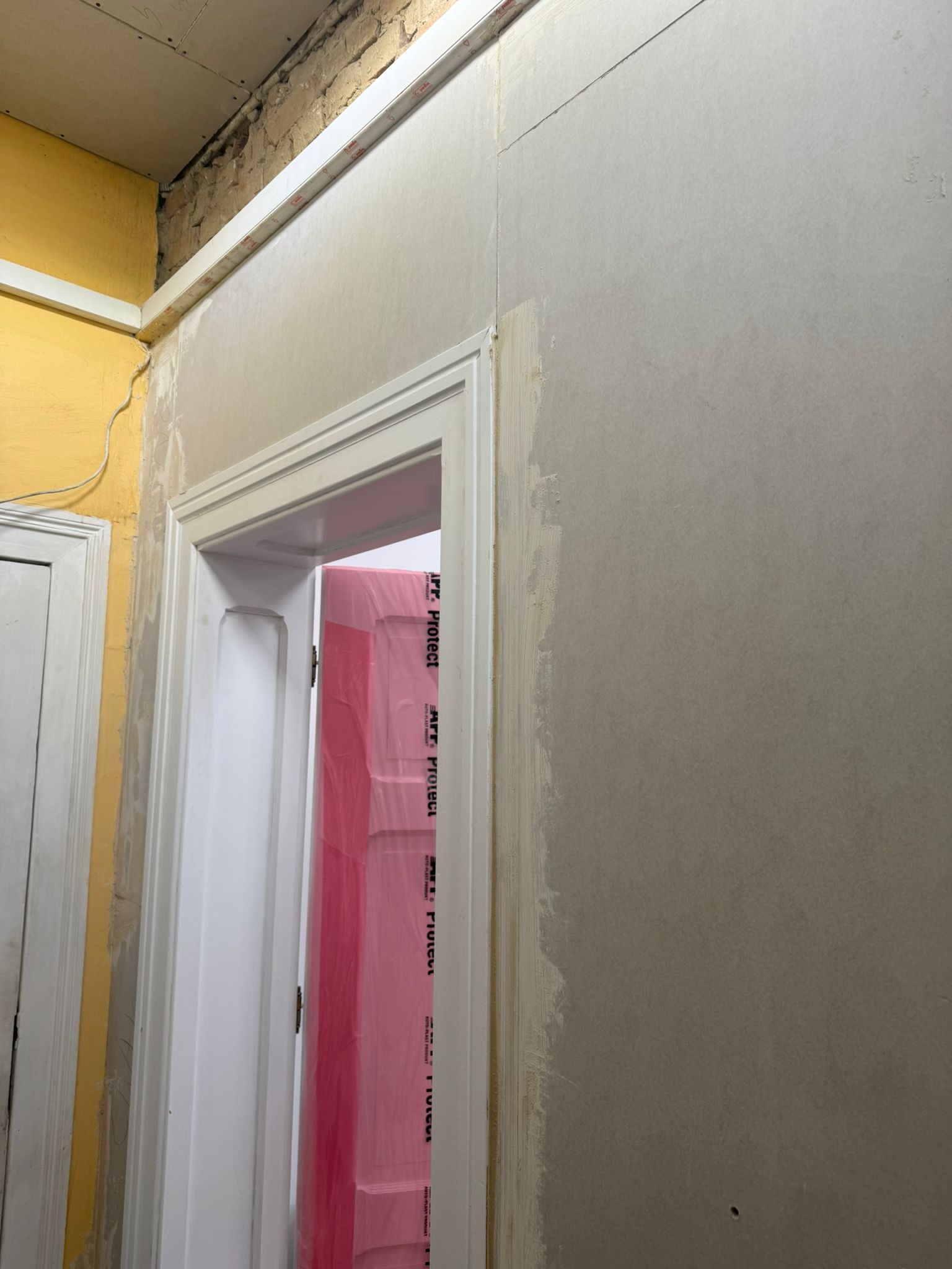 Ściana z płyt gipsowo-kartonowych w trakcie szpachlowania, widoczne zabezpieczone folią drzwi. Prace wykończeniowe wewnątrz pomieszczenia.