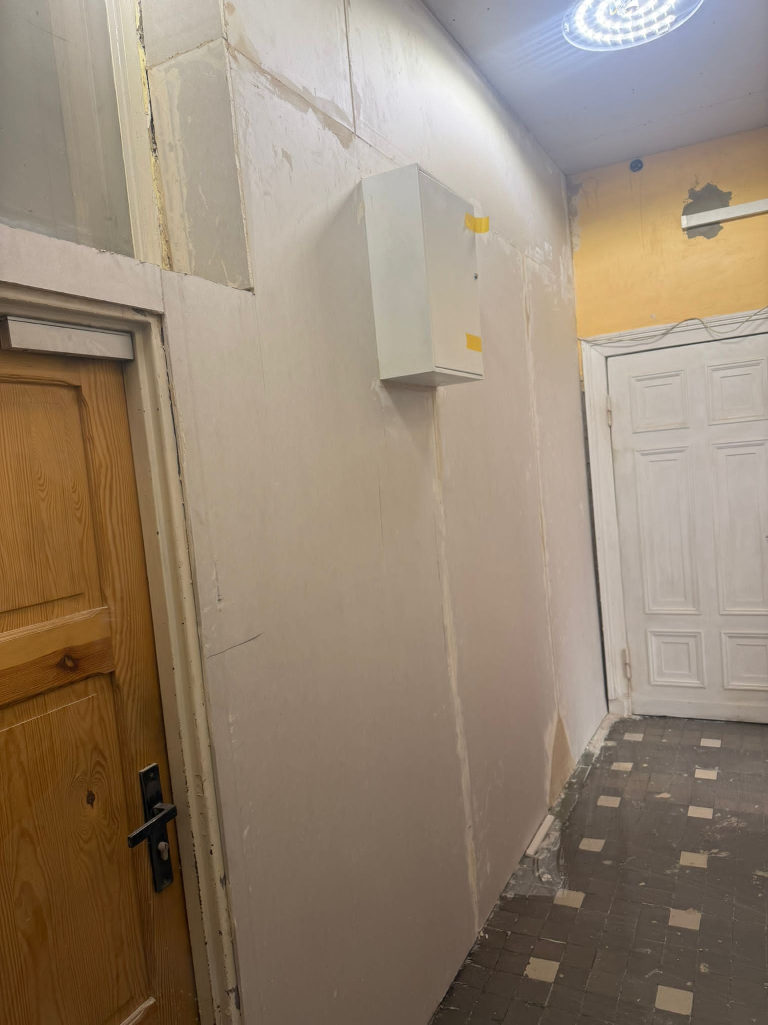 Ściana po szpachlowaniu z widoczną puszką elektryczną, przygotowana do malowania. Fragment drewnianych drzwi po lewej stronie i drzwi wejściowe w tle.