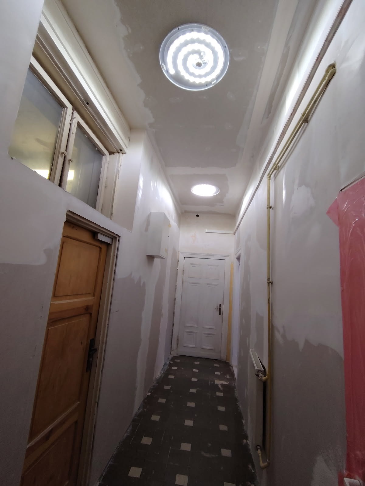 Korytarz w trakcie prac wykończeniowych: ściany przygotowane do malowania, widoczne szpachlowanie, sufit z oświetleniem. Podłoga z ciemną, kwadratową mozaiką.