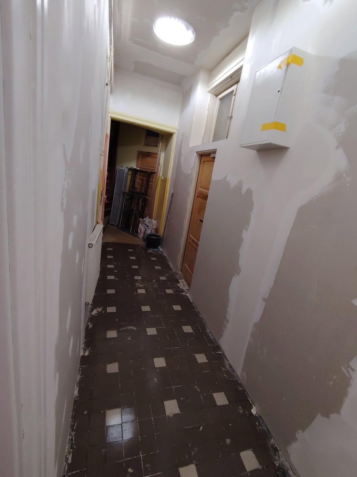 Korytarz w trakcie szpachlowania ścian, widoczne ślady po taśmie malarskiej, brązowa podłoga z kwadratowych płytek. W tle widoczne drzwi i elementy wyposażenia.