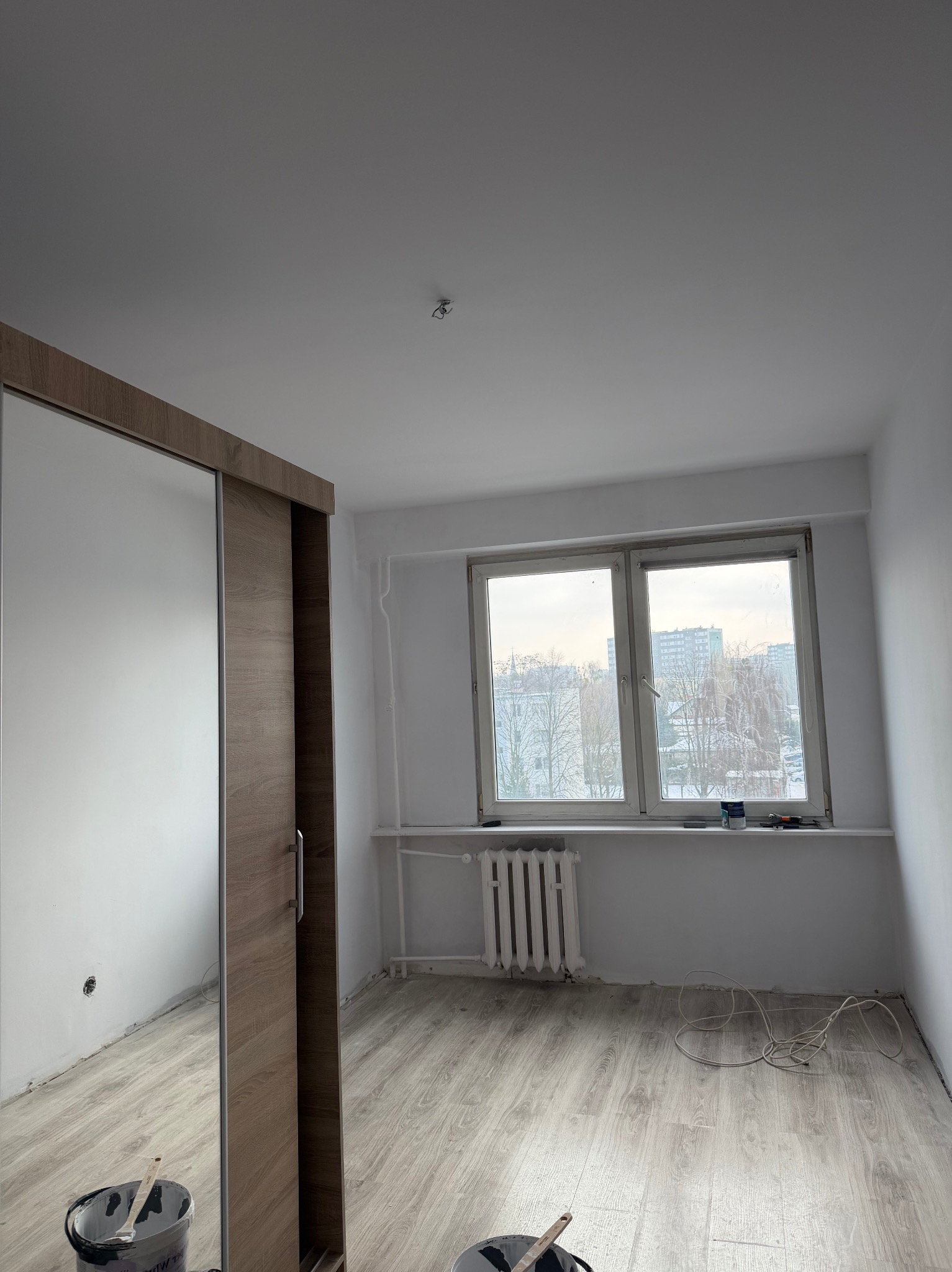 Pokój po malowaniu ścian na biało, z drewnianą podłogą i szafą z lustrem. Widoczne narzędzia malarskie i grzejnik pod oknem z widokiem na miasto.