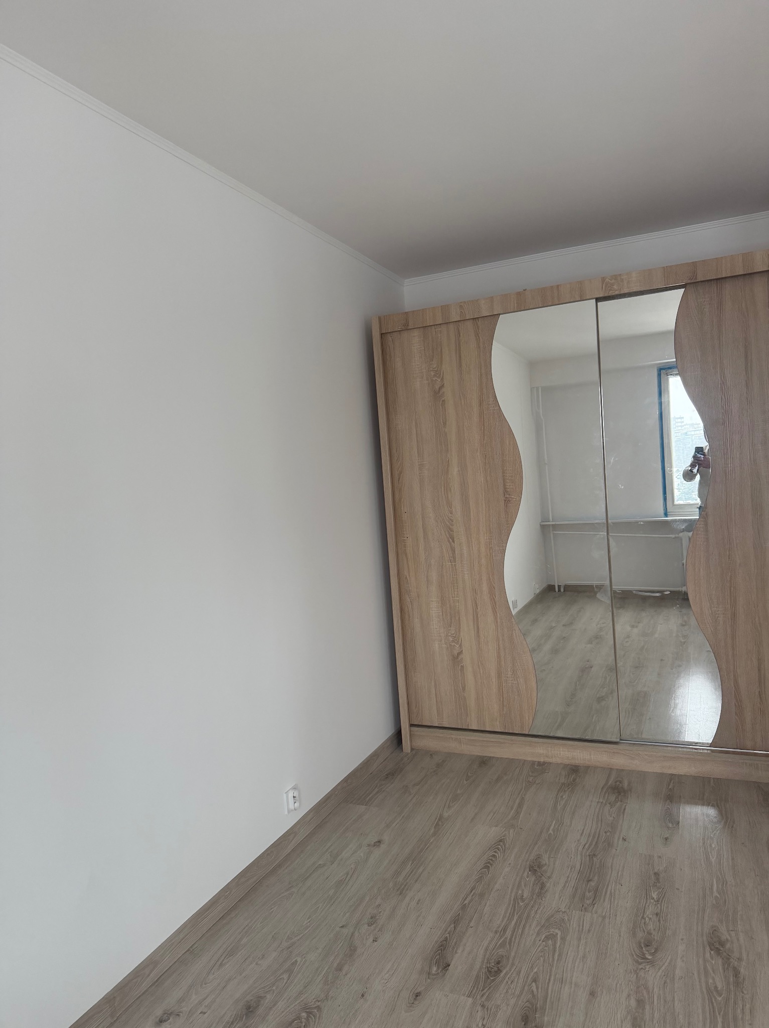 Jasne panele podłogowe w pokoju z białą ścianą i szafą z lustrzanymi drzwiami przesuwnymi o falistym kształcie. Widoczne gniazdko elektryczne.