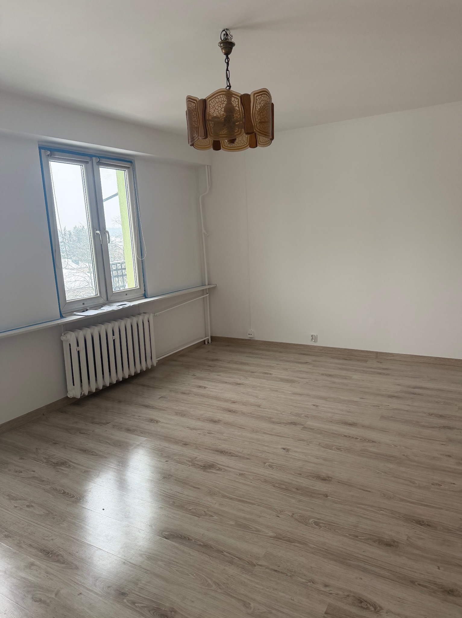 Jasne wnętrze pokoju po remoncie: panele podłogowe, białe ściany, okno z grzejnikiem i lampa w stylu retro. Widok na odnowione pomieszczenie.