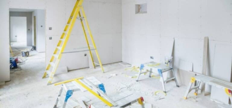 Wnętrze w trakcie prac wykończeniowych: ściany pokryte płytami gipsowo-kartonowymi, żółta drabina oparta o ścianę, widoczne narzędzia i materiały budowlane.