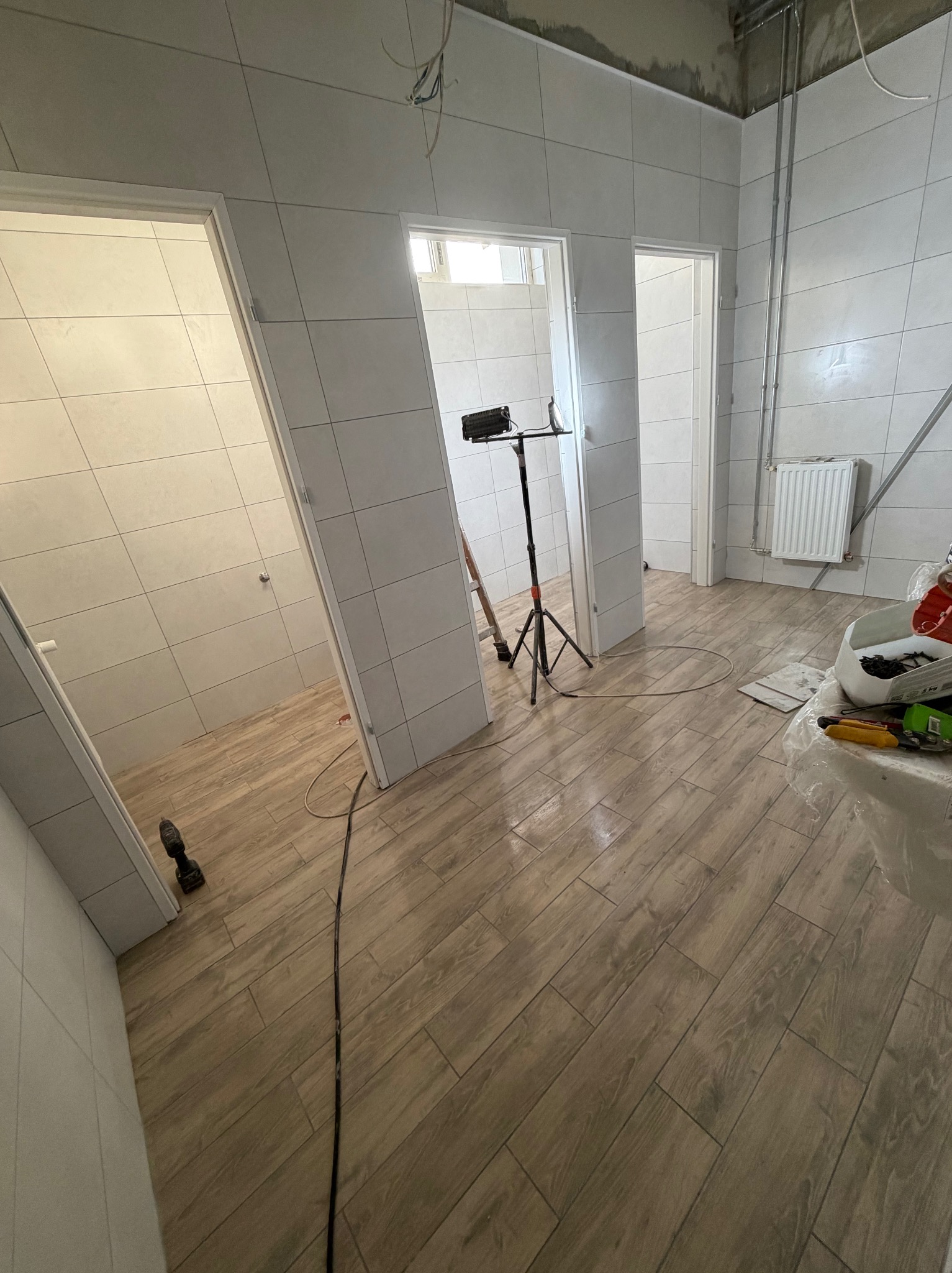 Wnętrze łazienki w trakcie remontu: położone płytki imitujące drewno na podłodze, białe płytki na ścianach, widoczne narzędzia i oświetlenie budowlane.