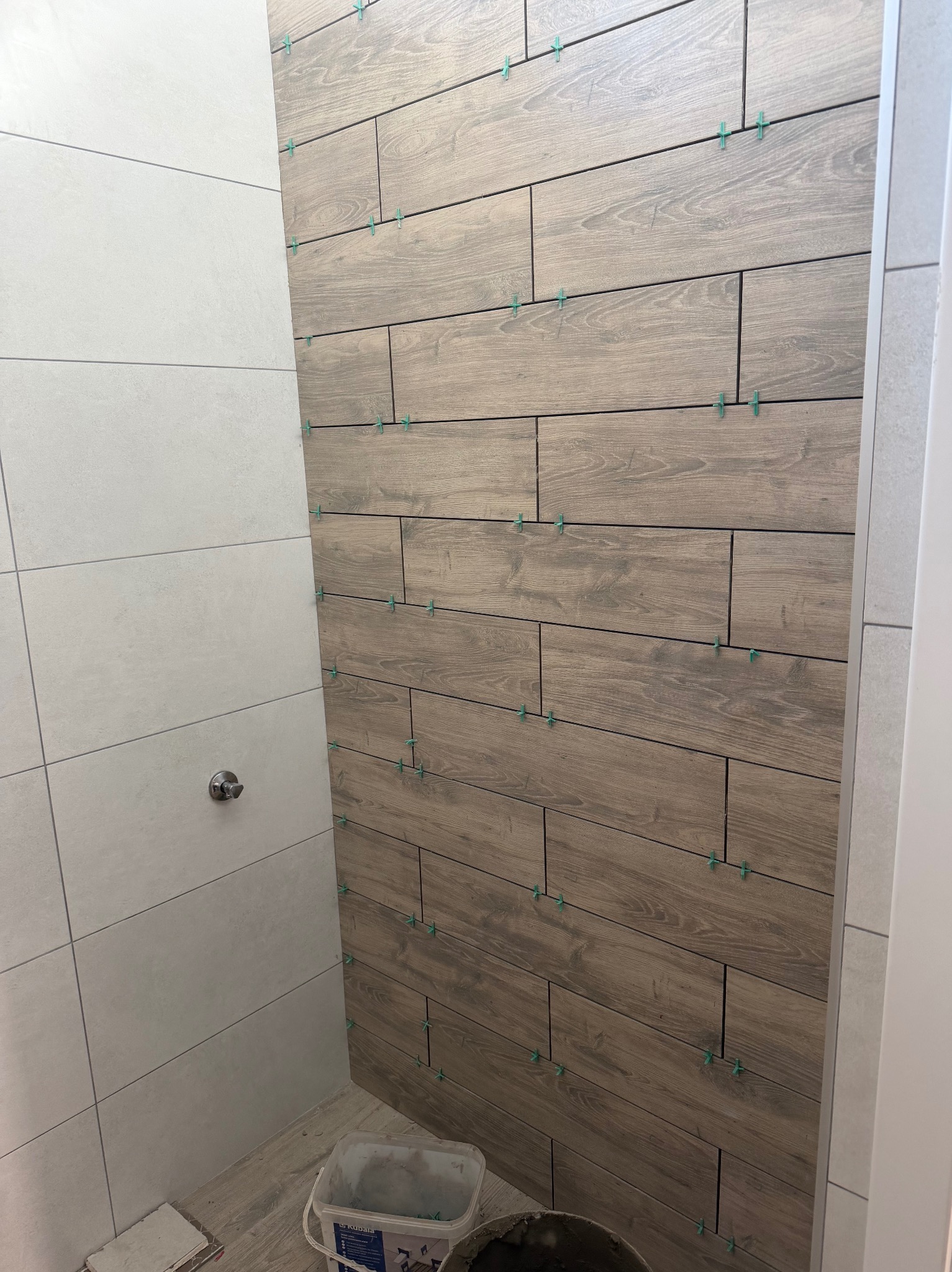 Ściana w trakcie układania płytek imitujących drewno, z widocznymi krzyżykami dystansowymi. Obok fragment ściany z jasnymi, prostokątnymi płytkami.