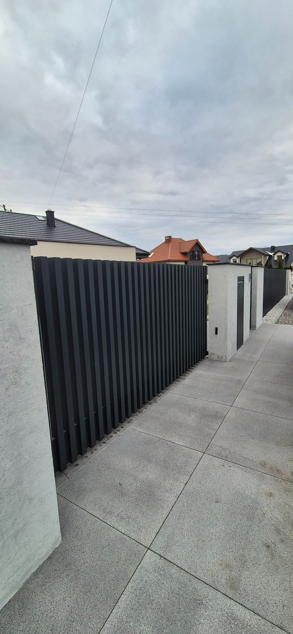 Nowoczesne ogrodzenie panelowe w kolorze grafitowym z betonowymi słupkami, widok z zewnątrz na tle domów jednorodzinnych i szarej kostki brukowej.