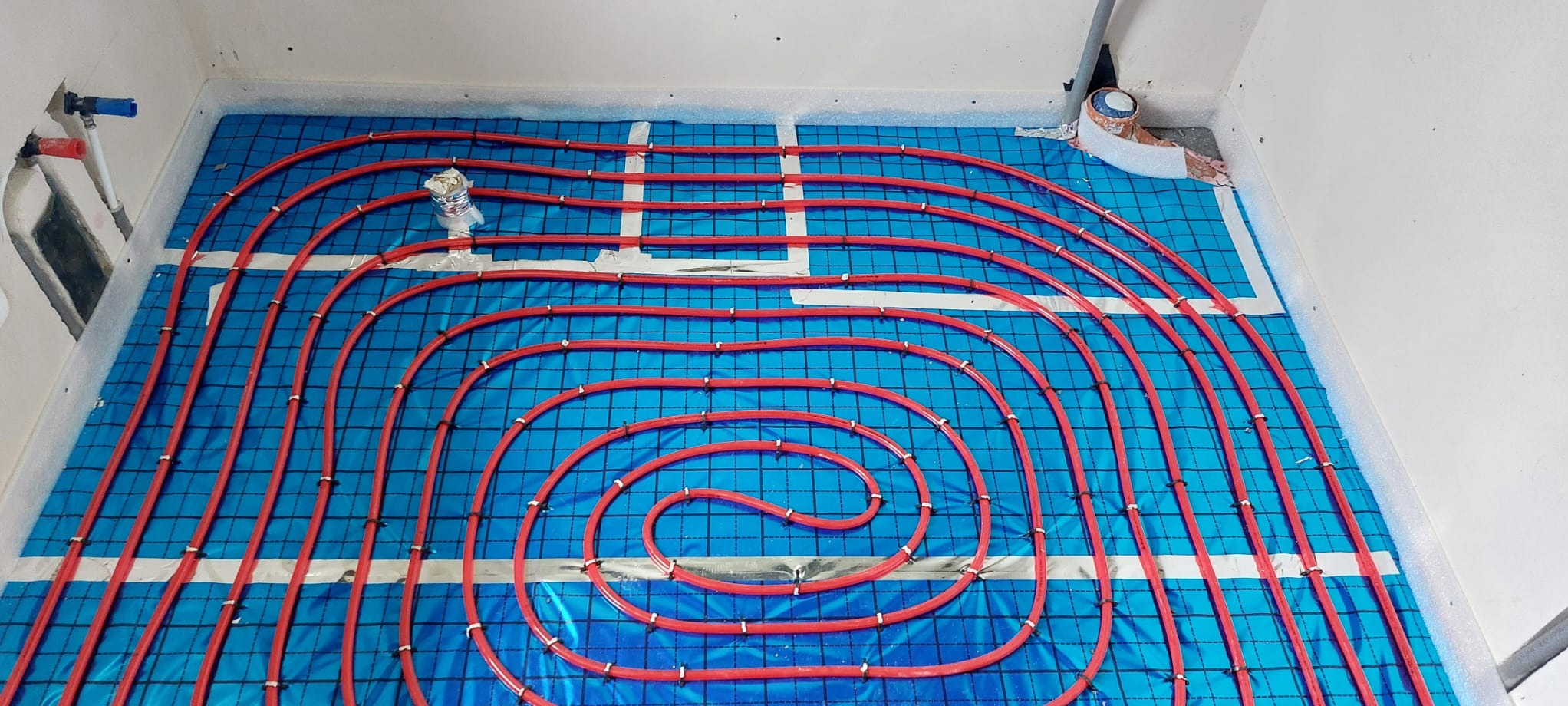 Czerwone rury ogrzewania podłogowego ułożone spiralnie na niebieskiej macie izolacyjnej z białymi paskami, widoczne elementy instalacji hydraulicznej przy ścianie.