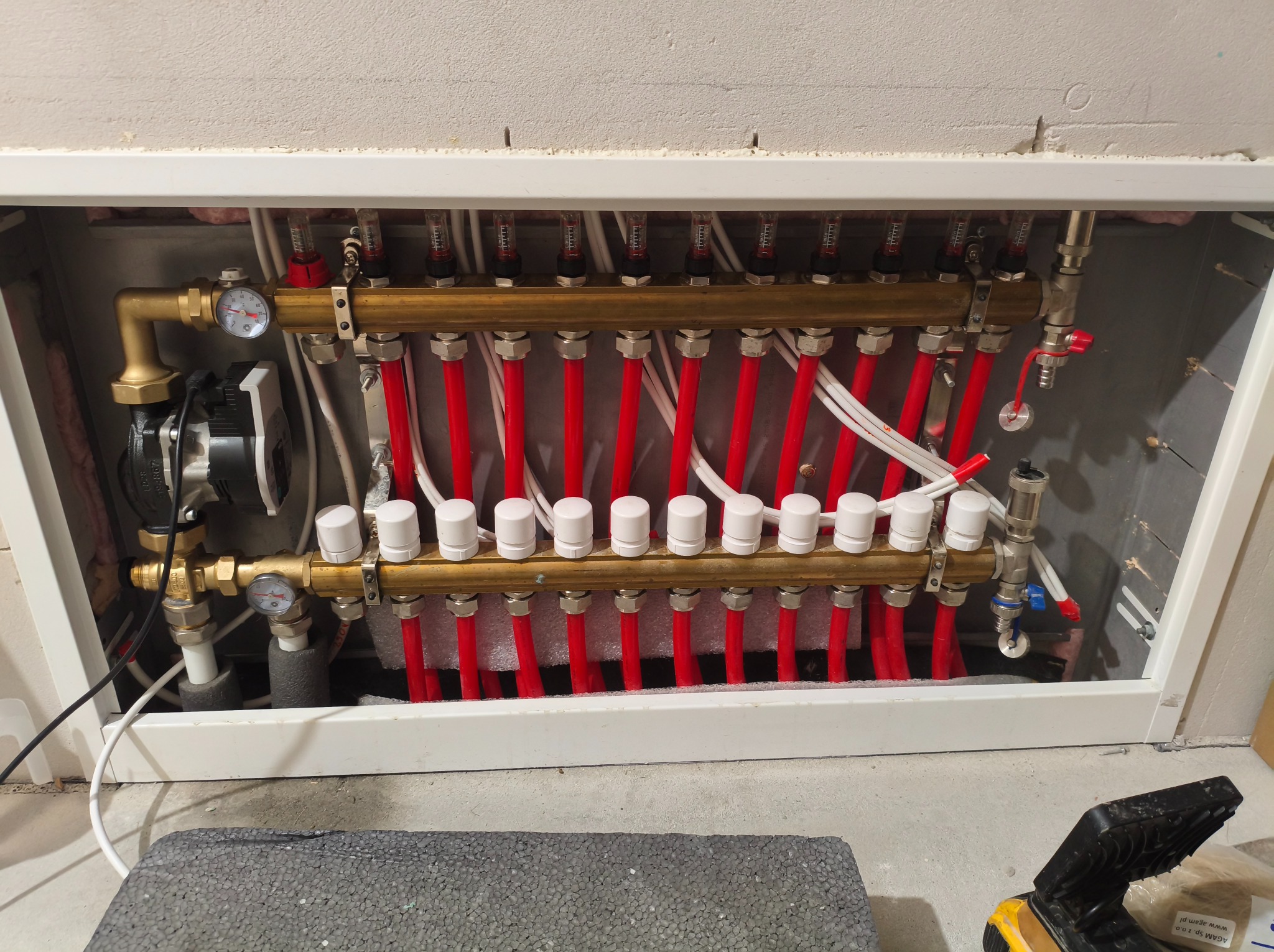 Rozdzielacz ogrzewania podłogowego z czerwonymi rurami, manometrami i białymi zaworami, umieszczony w białej wnęce w ścianie, widok z dołu.