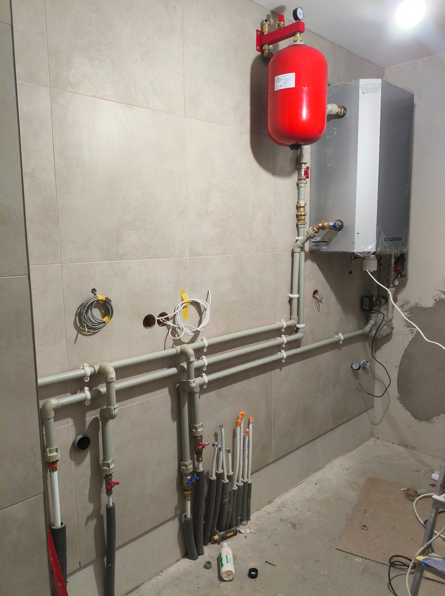 Instalacja grzewcza w kotłowni: czerwony zbiornik wyrównawczy, piec Stalmark, rury z zaworami, kable. Widoczne narzędzia i materiały instalacyjne.