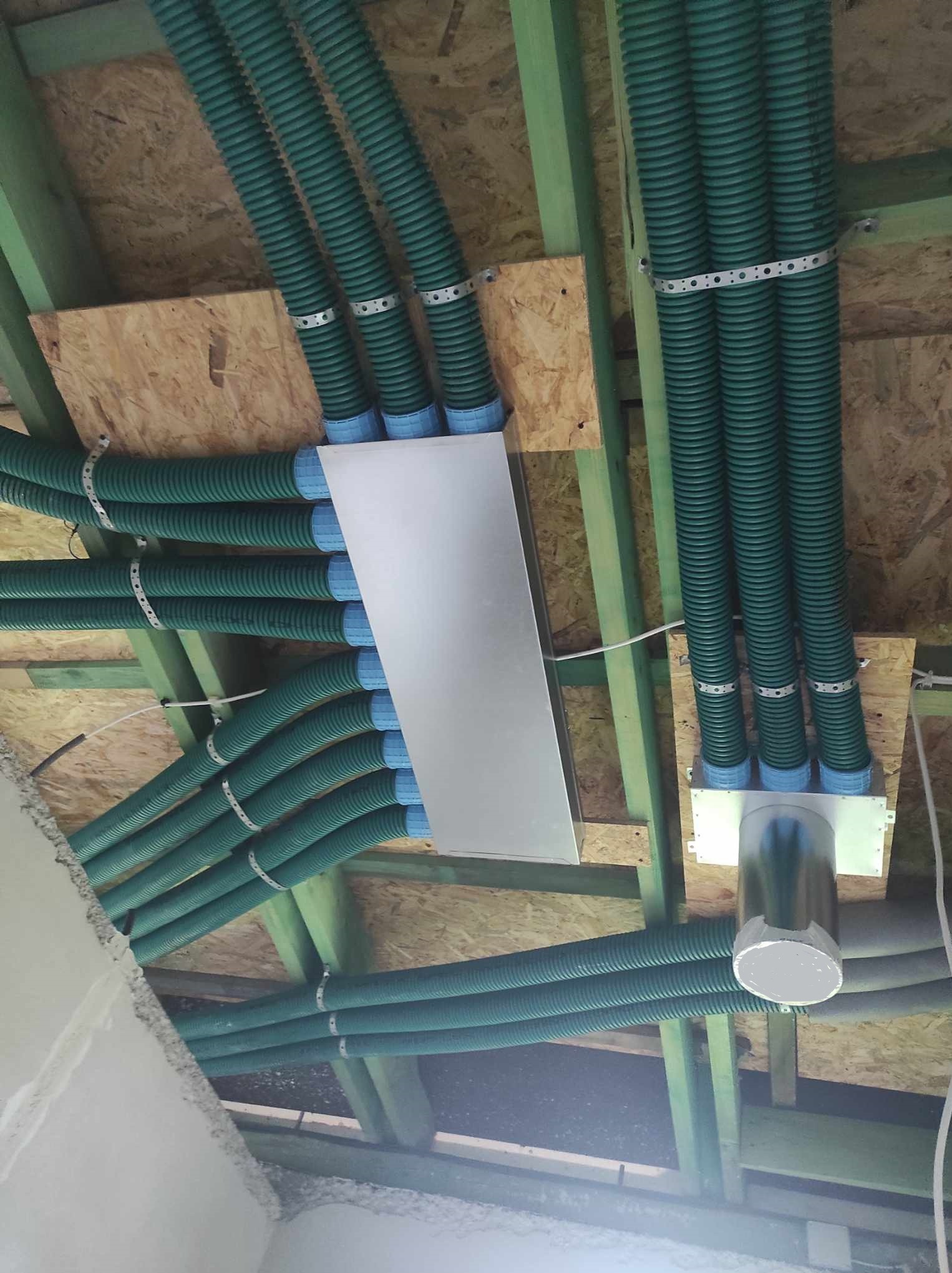 Zielone, karbowane rury wentylacyjne zamontowane pod drewnianym stropem, zabezpieczone metalowymi obejmami. Widoczne elementy mocujące i płyta montażowa.