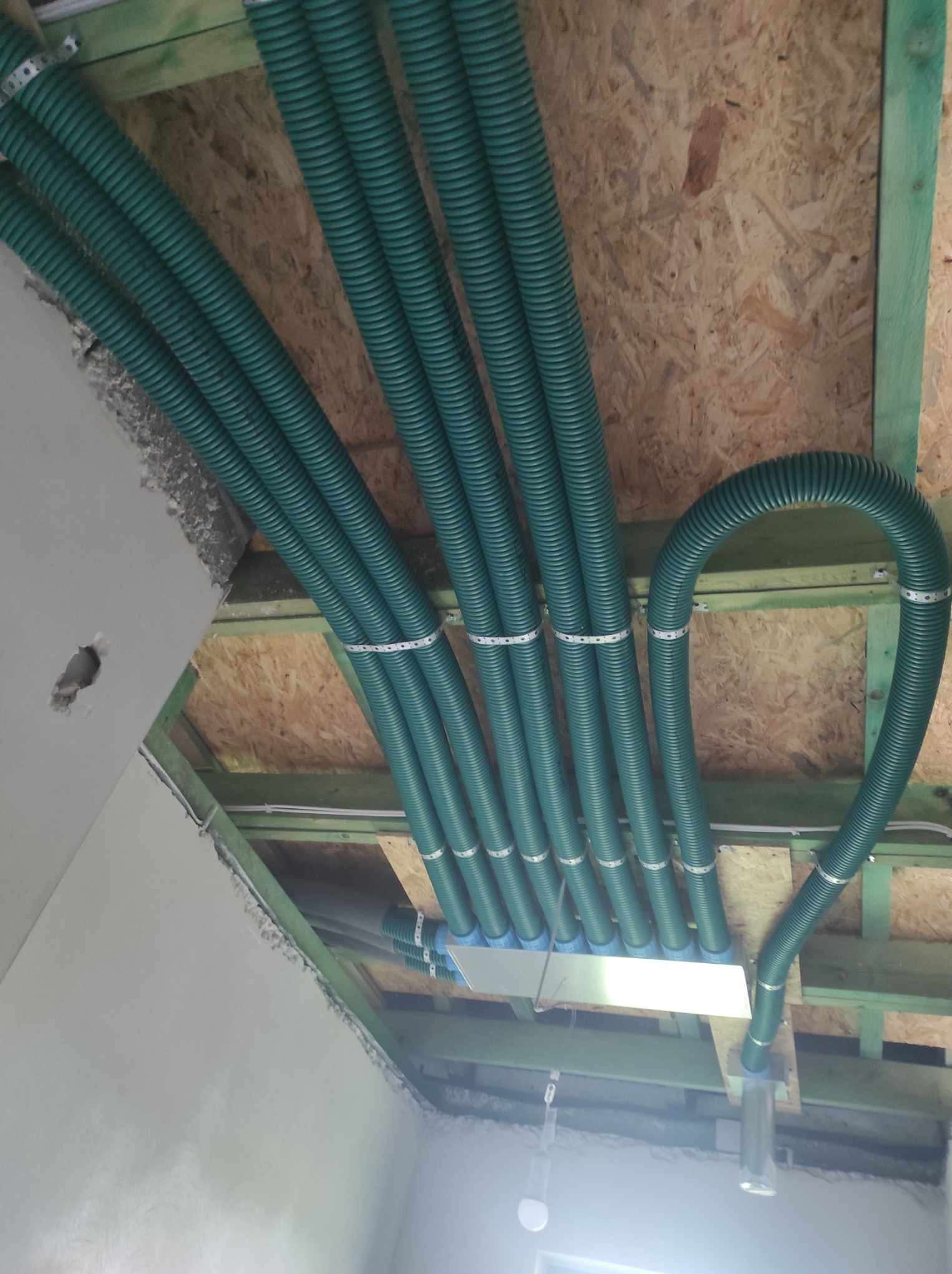 Zielone, karbowane rury wentylacyjne zamontowane pod drewnianym stropem, zabezpieczone metalowymi opaskami. Widoczne elementy instalacji i surowy stan wykończenia.