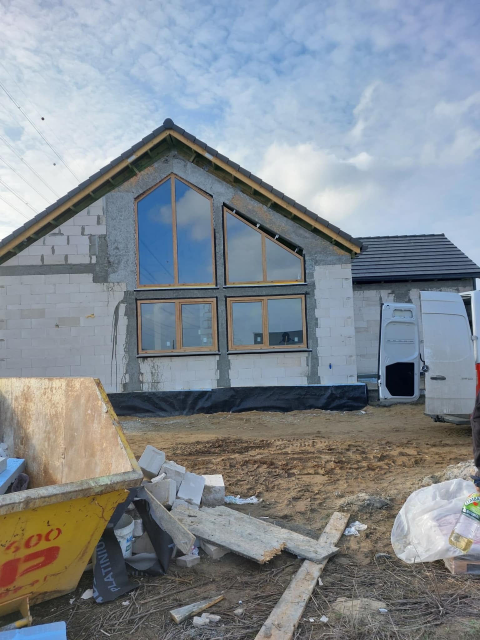 Surowy stan domu z zamontowanymi oknami o drewnianych ramach, widoczny kontener na gruz i częściowo samochód dostawczy. Budynek w trakcie budowy, z widocznymi bloczkami.