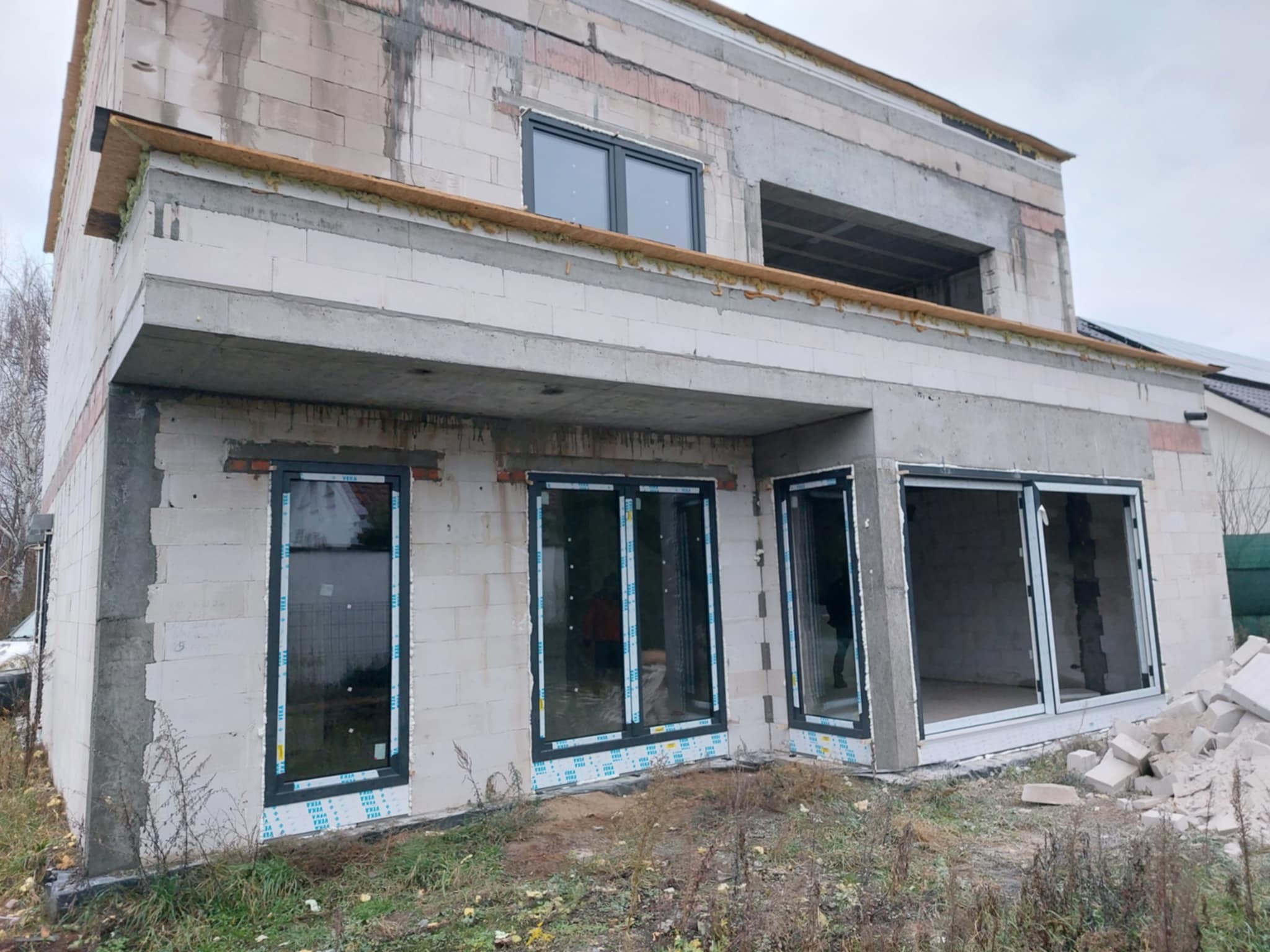 Surowy stan domu w budowie z zamontowanymi oknami PCV w grafitowych ramach. Widoczne bloczki, betonowe nadproża i przygotowane otwory okienne.