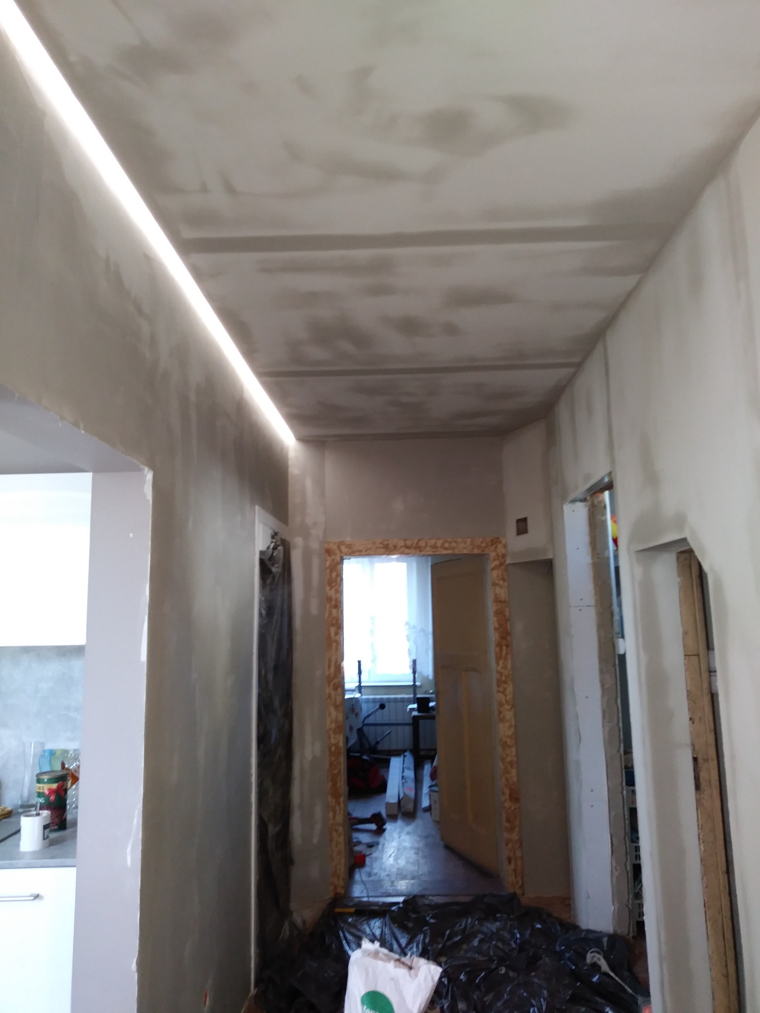 Korytarz w trakcie prac wykończeniowych: szpachlowane ściany i sufit z oświetleniem LED, widoczne przygotowania do dalszych prac. Folia ochronna na podłodze.