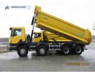 Żółta wywrotka z podniesioną skrzynią ładunkową, widoczna z boku na utwardzonym placu. Na skrzyni zielone napisy, w tle drzewa i pochmurne niebo.