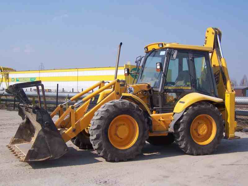 Żółta koparko-ładowarka JCB 4CX z łyżką z przodu i ramieniem kopiącym z tyłu, stojąca na placu budowy z widocznym budynkiem w tle.