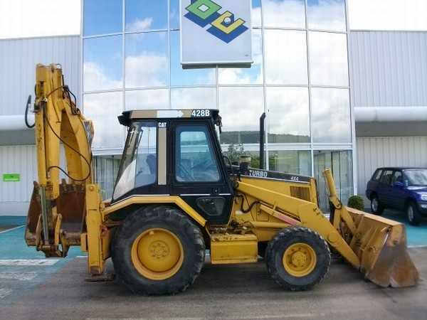 Żółta koparko-ładowarka CAT 428B Turbo zaparkowana przed nowoczesnym budynkiem z przeszkloną fasadą i niebieskim samochodem w tle.