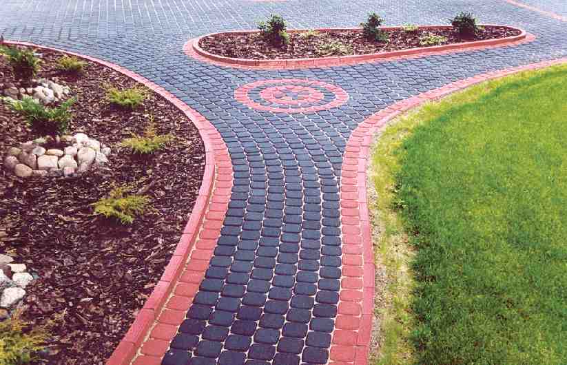 Czarno-czerwona kostka brukowa ułożona w kształt rozwidlającej się ścieżki z okrągłym wzorem pośrodku, obrzeża z czerwonej kostki, trawnik i rabaty z korą i roślinami po bokach.