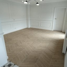 Paweł Albert Twój Kwadrat - Jasne, przestronne wnętrze z parkietem ułożonym we wzór jodełki. Białe ściany zdobione sztukaterią, nowoczesna lampa sufitowa. Fragment podłogi z wzorem gwiazd na wejściu.