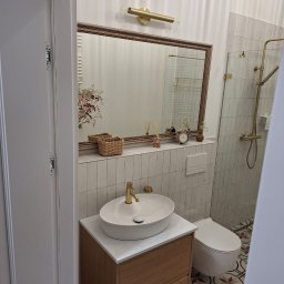 Paweł Albert Twój Kwadrat - Jasna łazienka po remoncie: białe płytki, umywalka na drewnianej szafce z marmurowym blatem, lustro w ozdobnej ramie, złota armatura i wzorzysta podłoga.