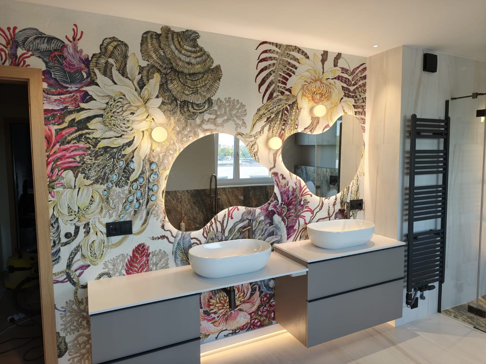Wykończona łazienka z umywalkami nablatowymi, podświetlanymi lustrami o nieregularnym kształcie i tapetą w kwiatowe wzory. Grzejnik drabinkowy na ścianie.