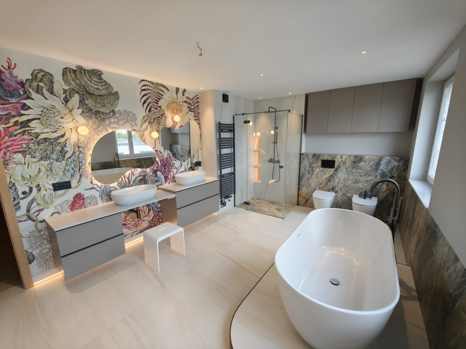 Nowoczesna łazienka z tapetą w kwiaty, owalnym lustrem z podświetleniem LED, wolnostojącą wanną i kabiną prysznicową. Wykończenie w jasnych barwach.