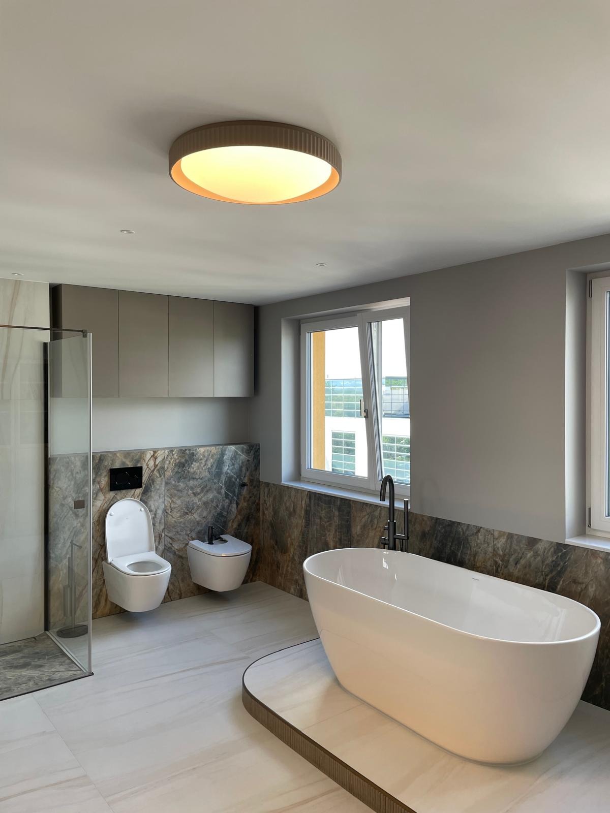 Nowoczesna łazienka z wolnostojącą wanną, marmurowymi akcentami i podwieszaną toaletą. Minimalistyczny design, jasne kolory i okrągła lampa sufitowa.