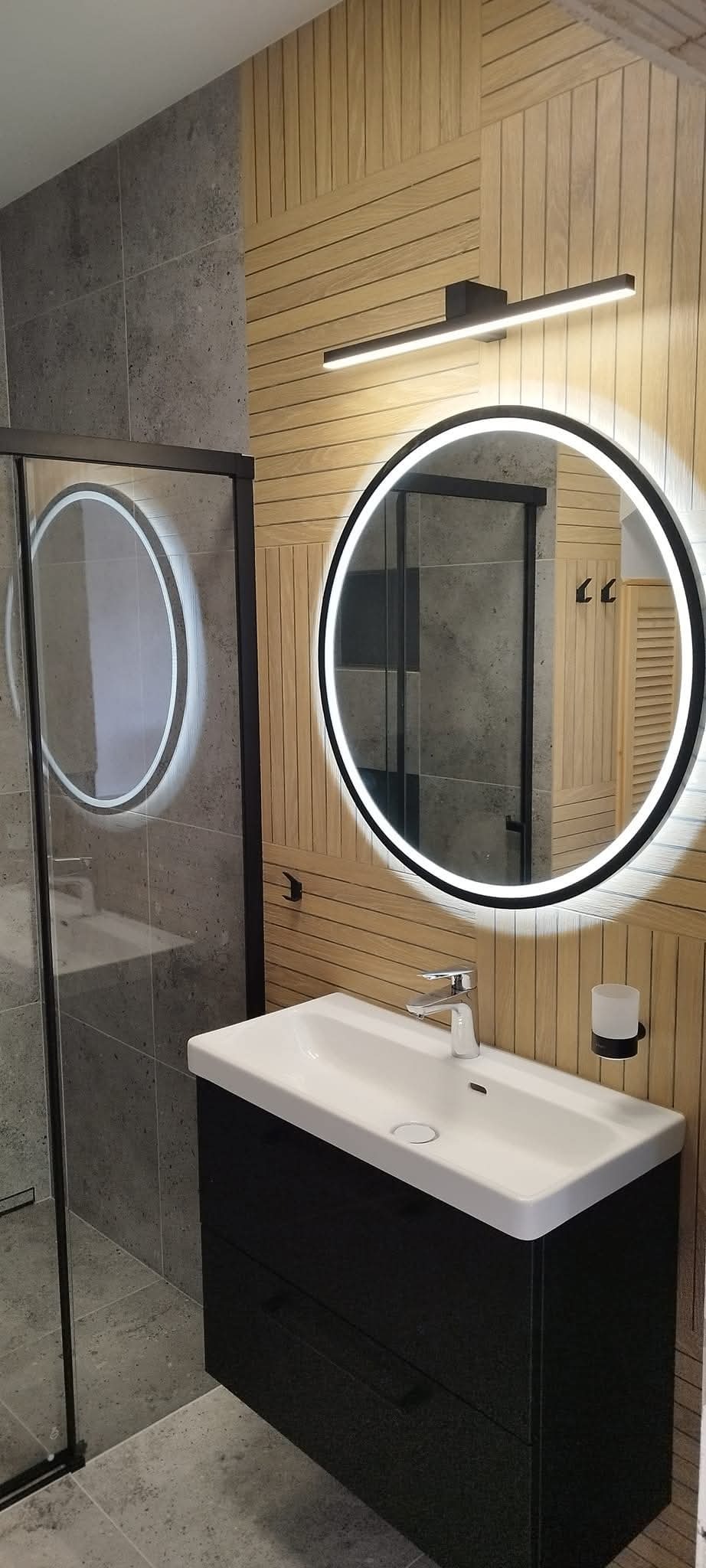 Nowoczesna łazienka z okrągłym lustrem LED, drewnianą boazerią i szarą glazurą. Umywalka nablatowa na czarnej szafce. Minimalistyczny design, ciepłe oświetlenie.