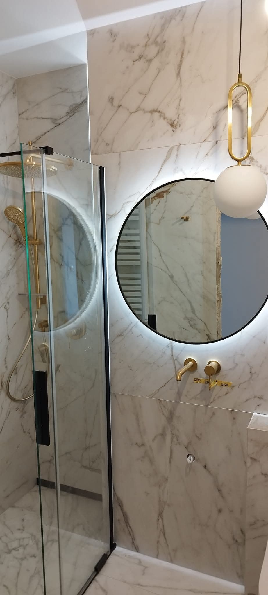 Elegancka łazienka z kabiną prysznicową, okrągłym lustrem z podświetleniem LED i złotą armaturą na tle marmurowych płytek. Nowoczesny design z dbałością o detale.