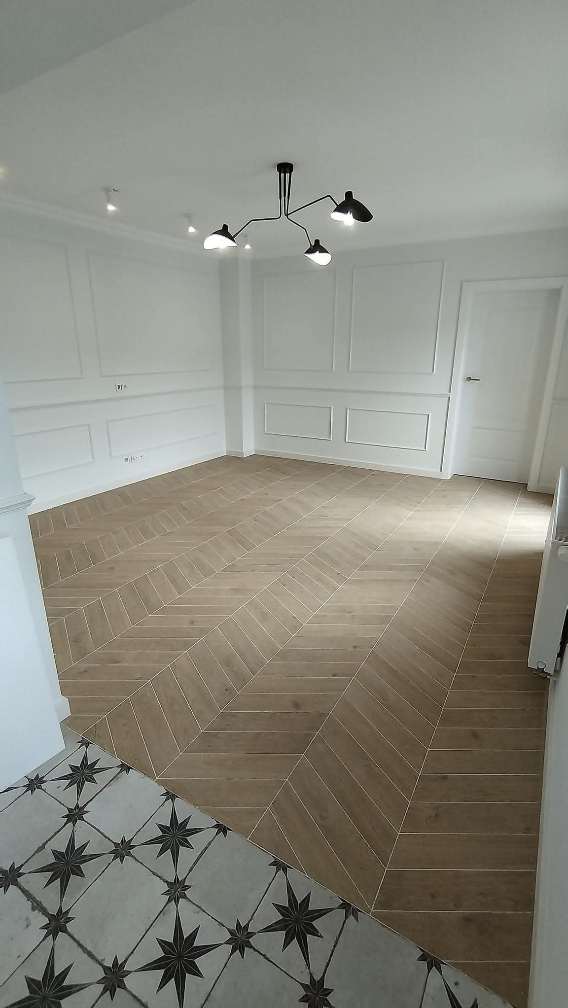 Jasne, przestronne wnętrze z parkietem ułożonym we wzór jodełki. Białe ściany zdobione sztukaterią, nowoczesna lampa sufitowa. Fragment podłogi z wzorem gwiazd na wejściu.