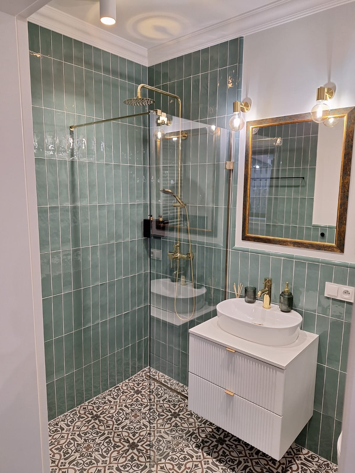 Wykończona łazienka z zielonymi, pionowo ułożonymi płytkami, złotą armaturą i wzorzystą podłogą. Umywalka nablatowa na białej szafce z frezowanymi frontami.