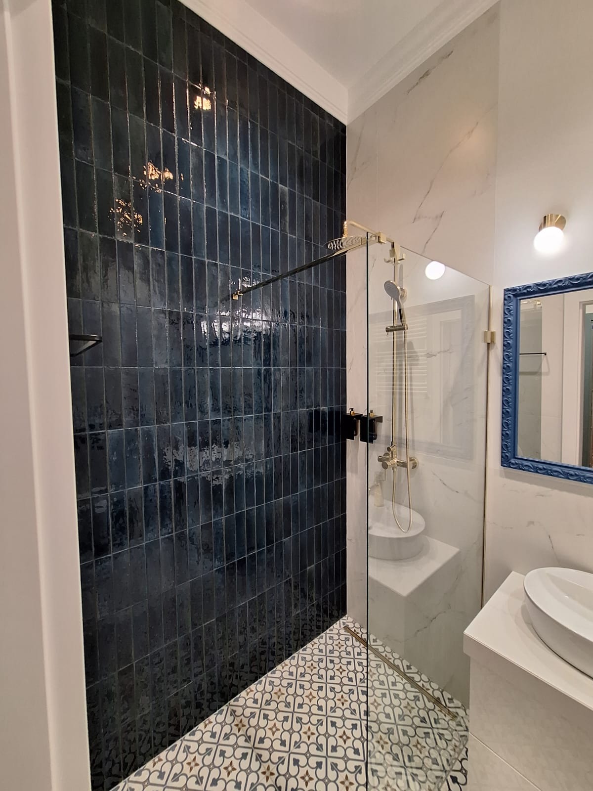 Elegancka łazienka po remoncie: ciemne, pionowe płytki na ścianie kabiny prysznicowej, wzorzysta podłoga i marmurowe wykończenia. Złote detale armatury.