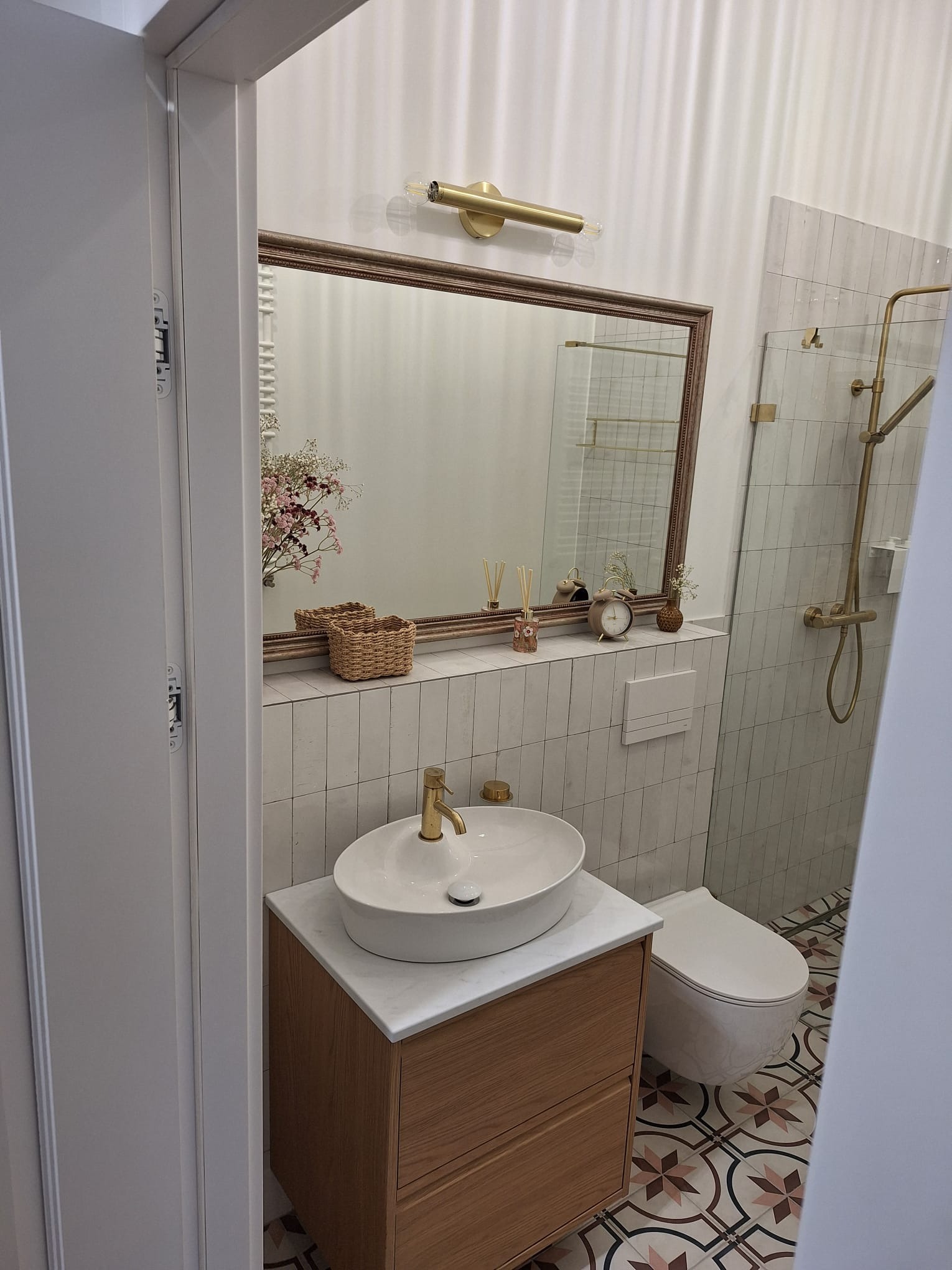 Jasna łazienka po remoncie: białe płytki, umywalka na drewnianej szafce z marmurowym blatem, lustro w ozdobnej ramie, złota armatura i wzorzysta podłoga.